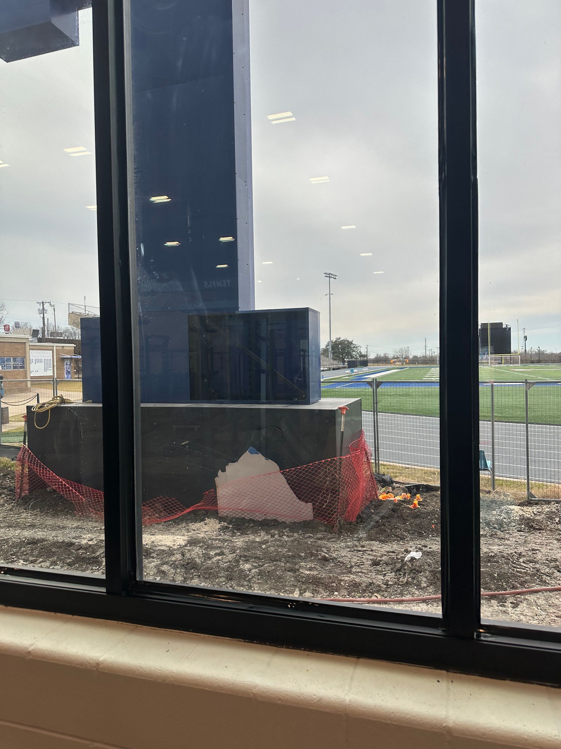 View through a window of a construction site with a blue structure, red fencing, and a cloudy sky.