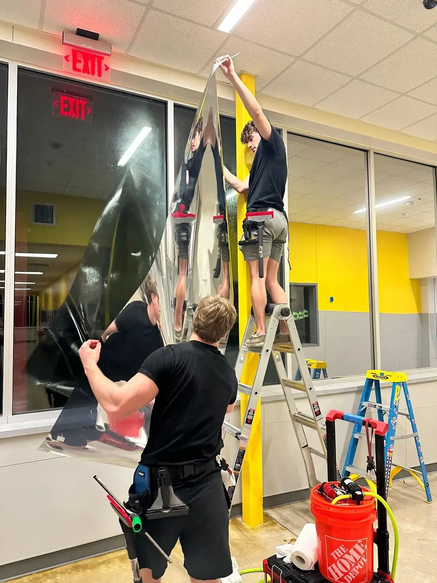 Three people installing reflective window tint in a school hallway. One on ladder, two on ground.