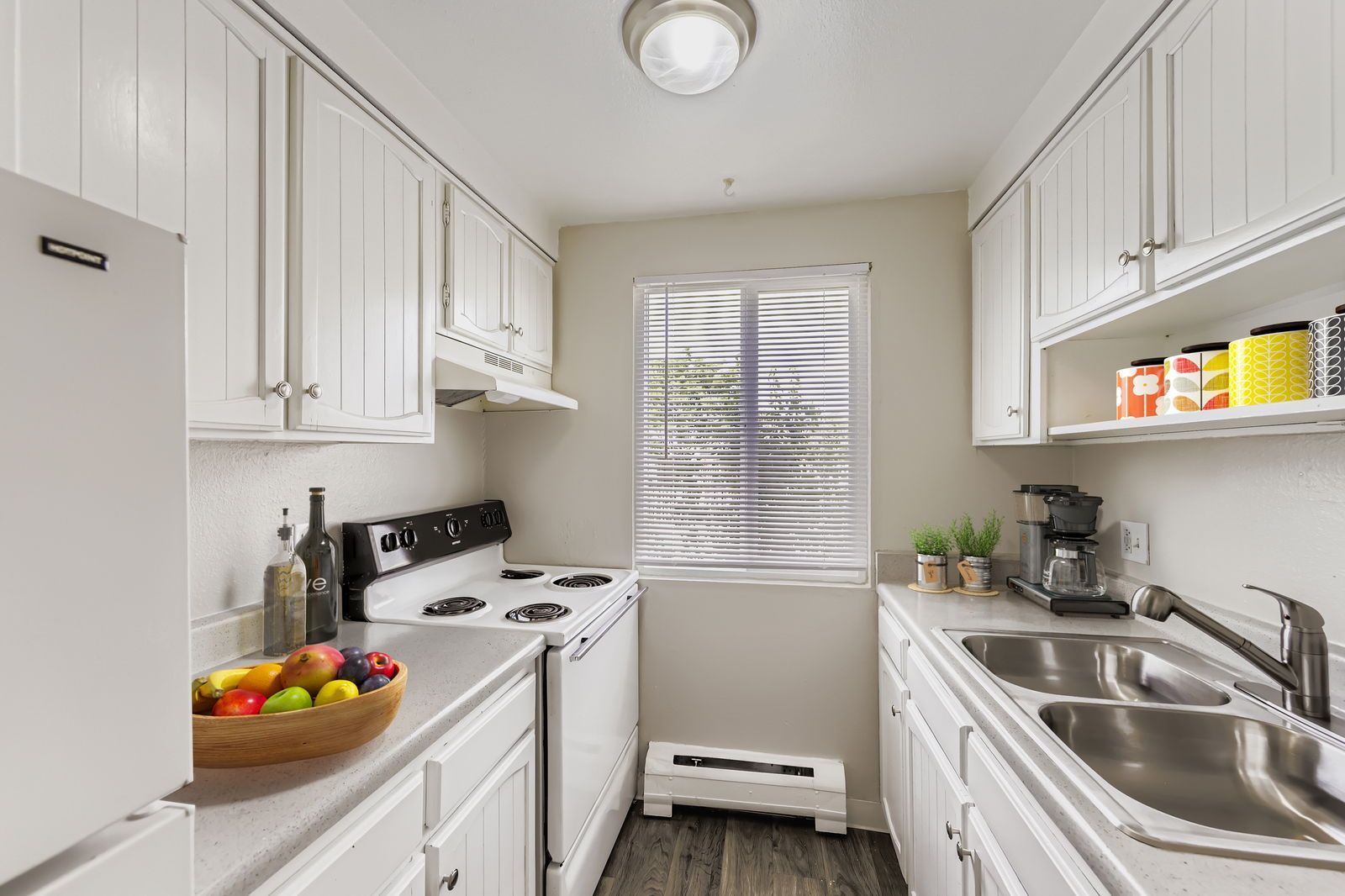 White galley kitchen with white cabinets, stove, fridge, double sink, and window with blinds.