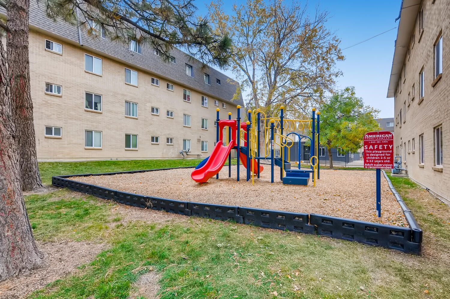 Playground with a red slide and blue-yellow climbing structures in a grassy courtyard between buildings.