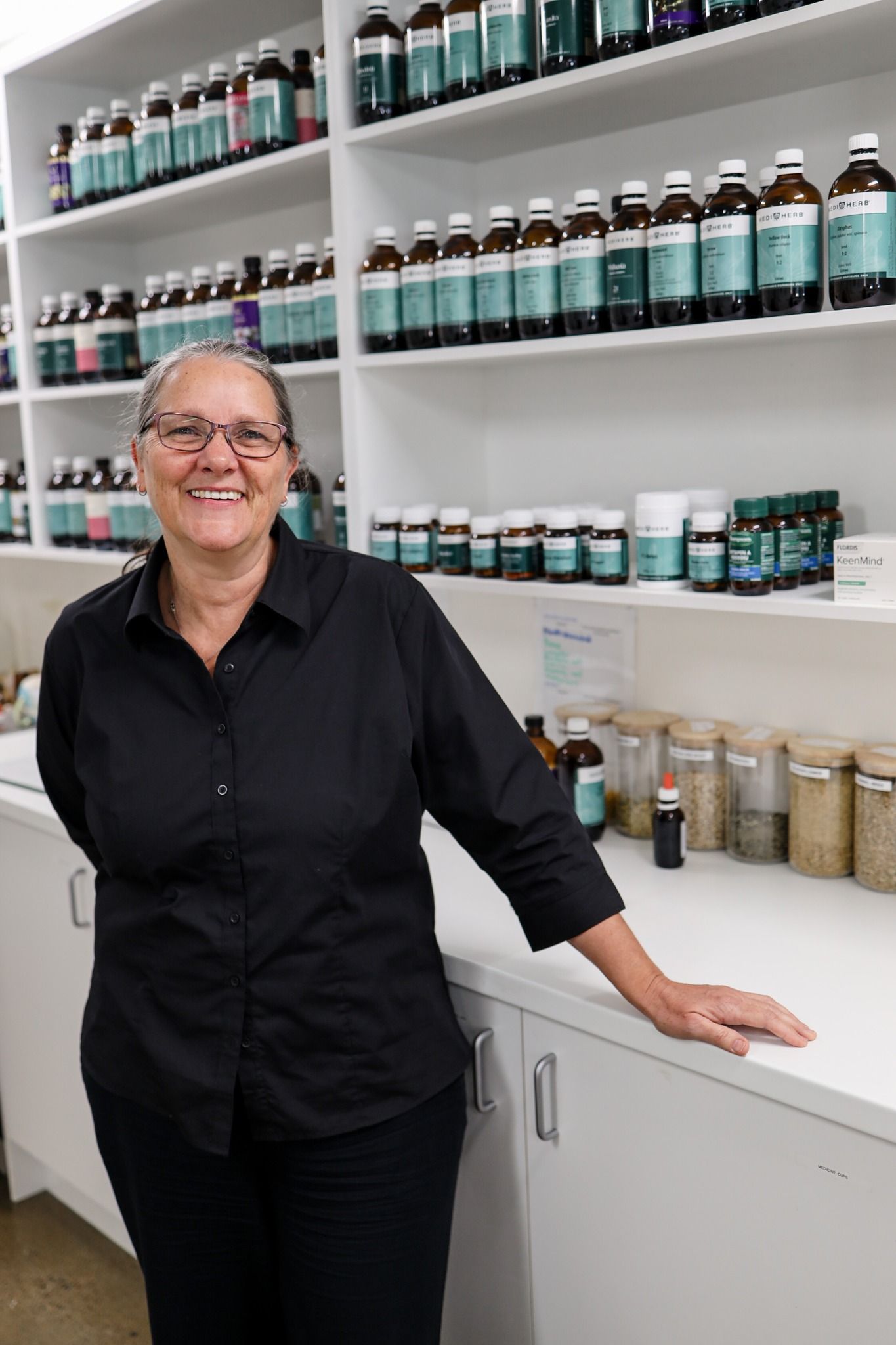 Woman in black shirt smiles in front of shelves filled with bottles in a store.