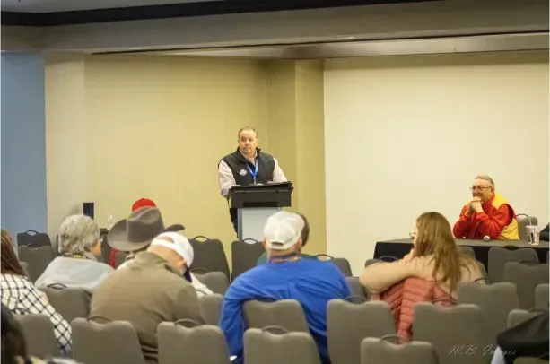 Man speaking at podium in front of a seated audience. Another man is seated at a table. Conference room.