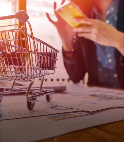 Small shopping cart beside papers while a person uses a tablet, with warm orange light in a workspace.