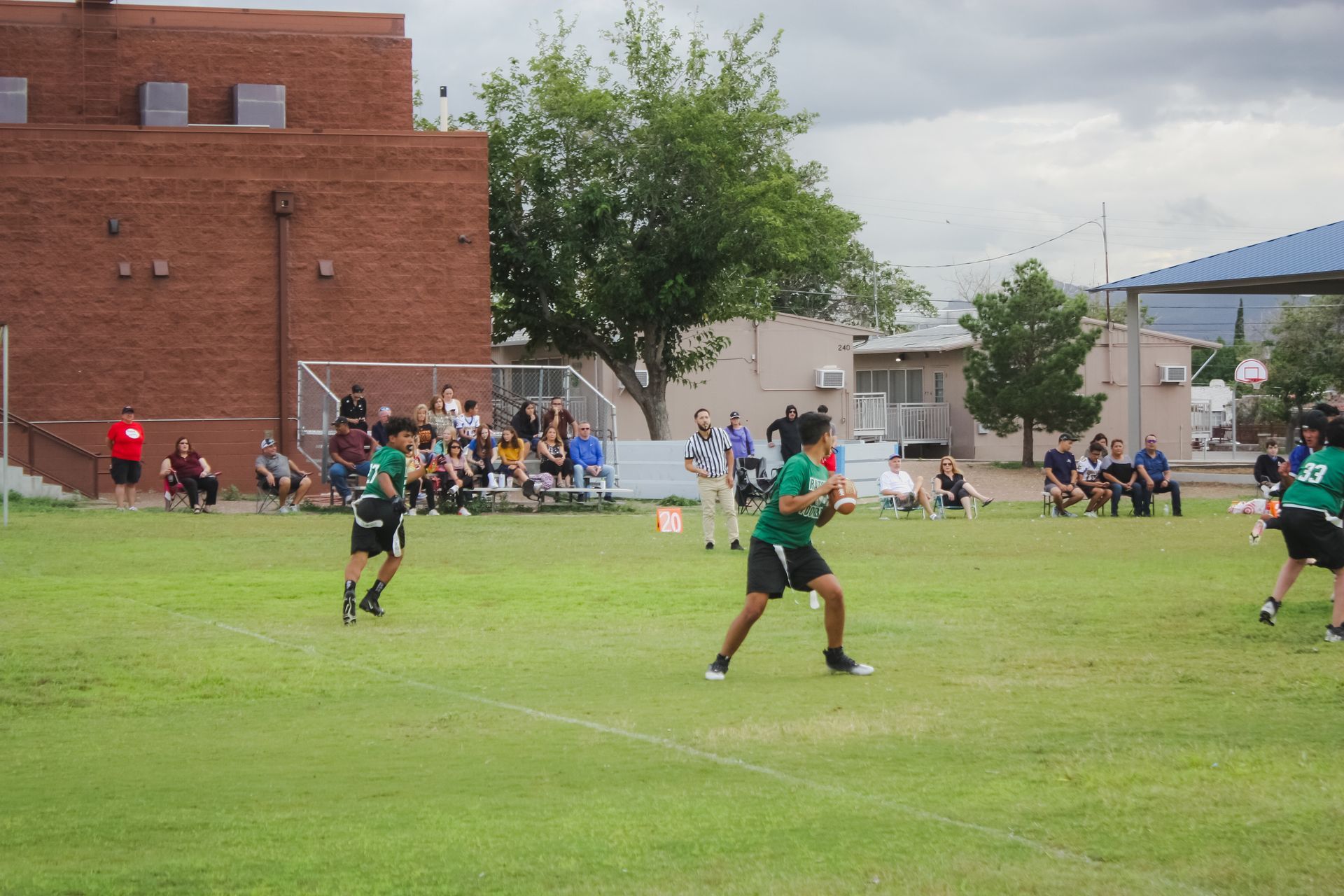 Girls playing flag football on a green field, spectators in bleachers, brick building in the background.