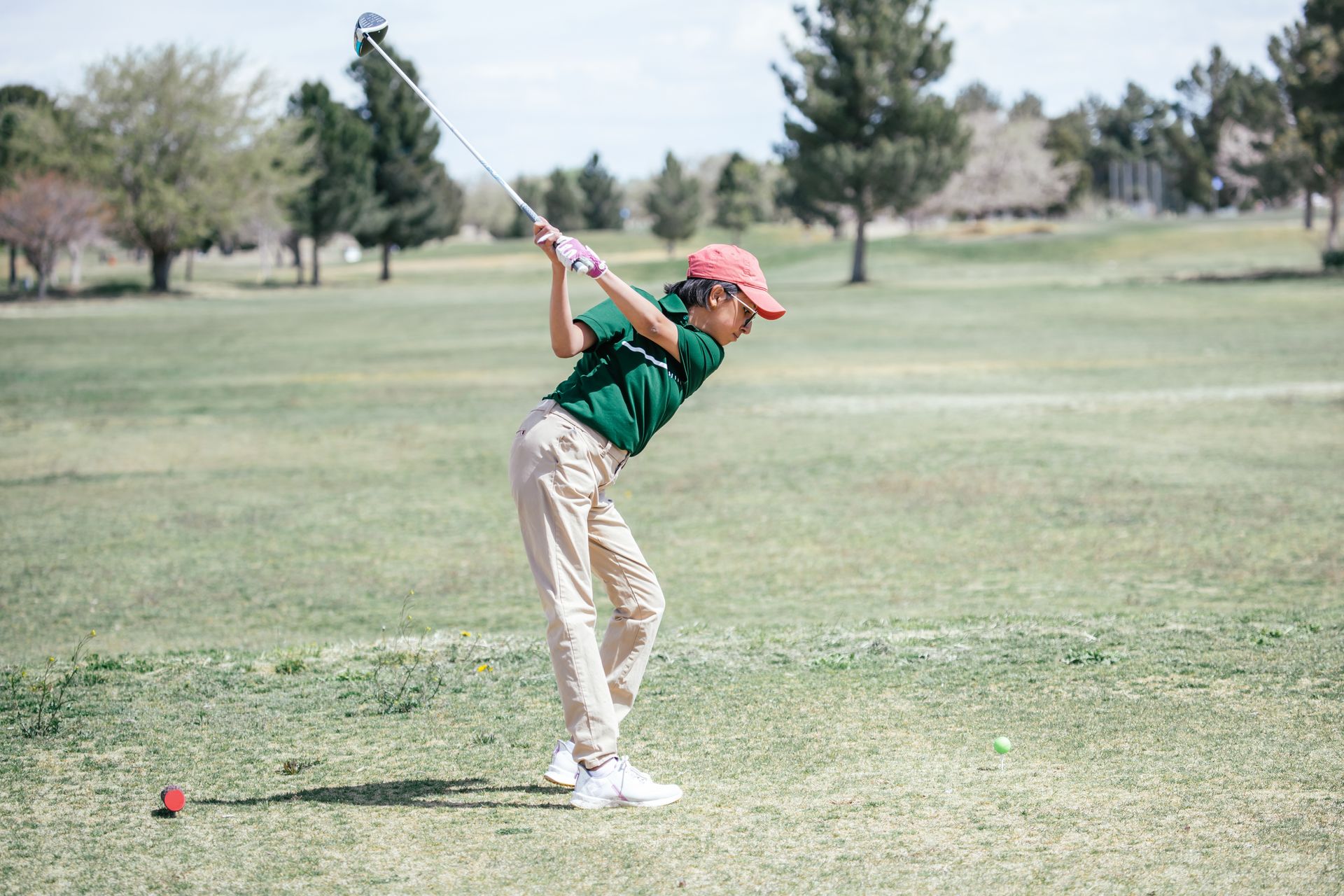 A young person in a green shirt and pink hat swings a golf club on a green golf course.