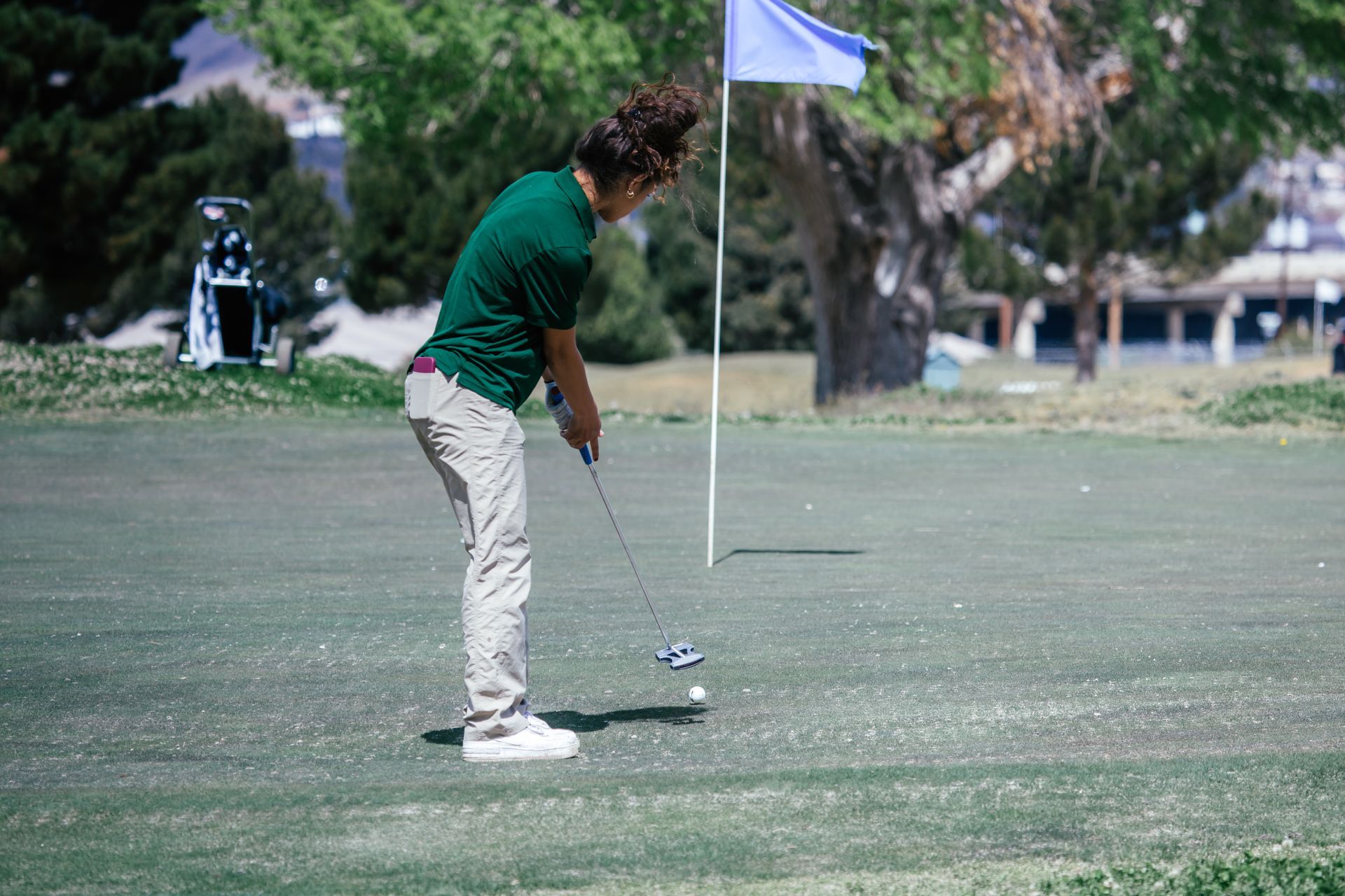 Golfer in green shirt and khakis putts on a green, blue flag behind him.