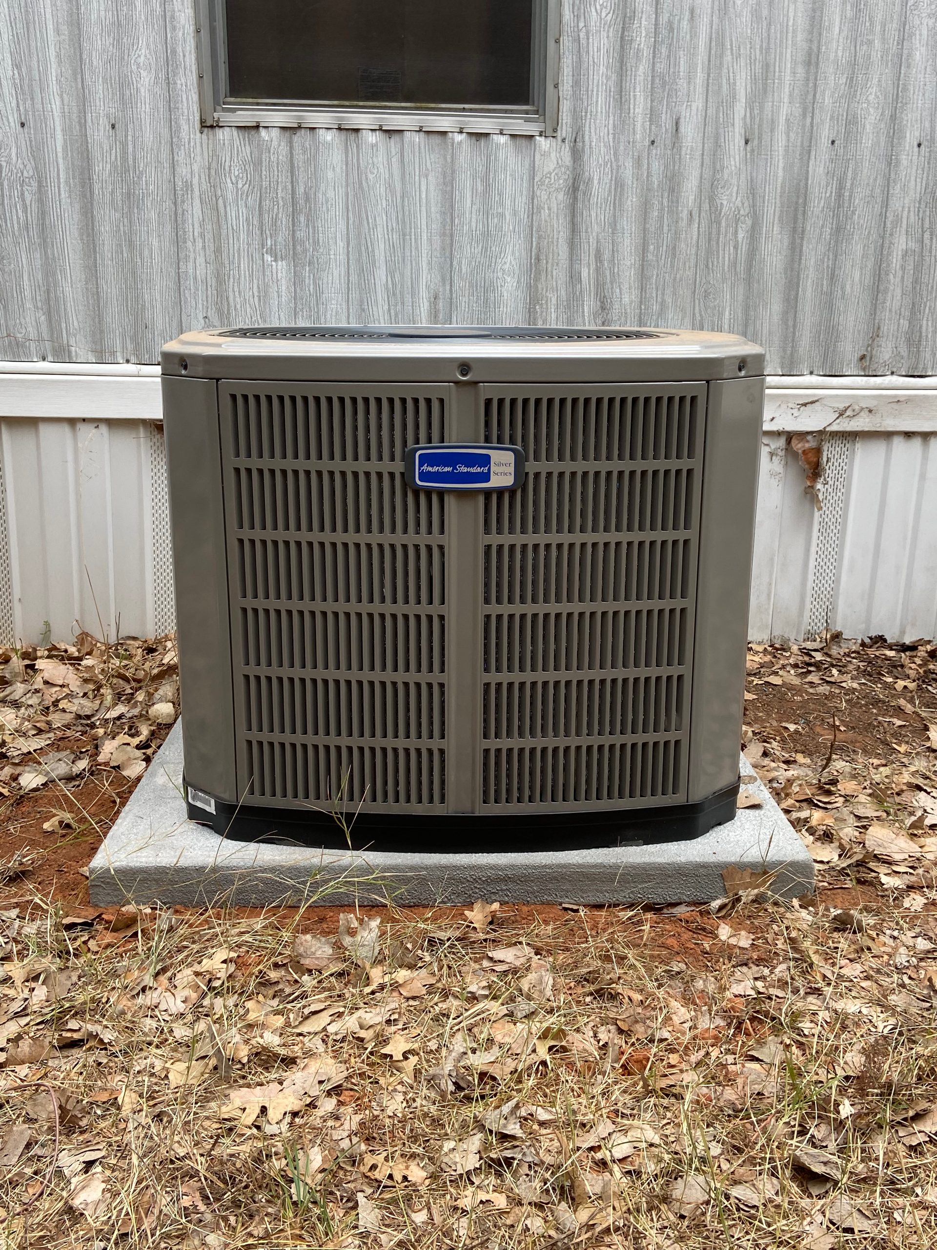 A gray air conditioner is sitting on a concrete platform in front of a house.