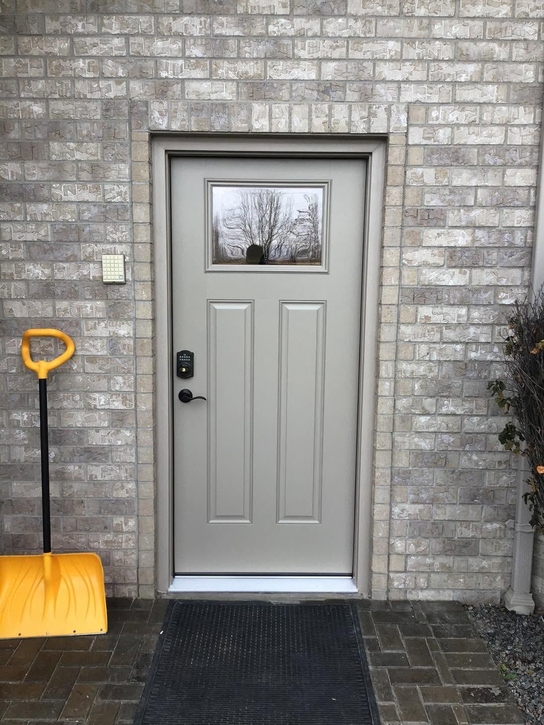 Front door of a gray brick house with a snow shovel leaning beside it and a mat on the steps