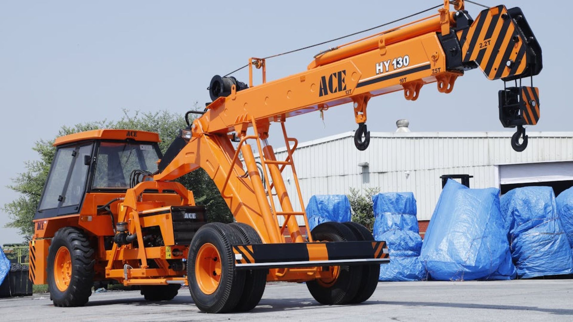 Grúa ACE Hydra 130 de color naranja sobre ruedas en un entorno al aire libre, al lado de un edificio y elementos cubiertos con una lona azul.