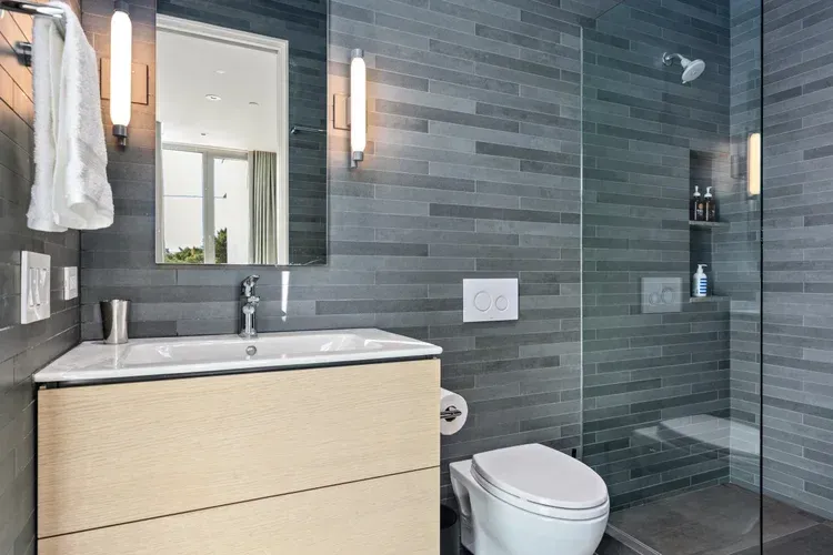 Modern bathroom with wood vanity, mirror, toilet, and glass shower in gray tile surroundings