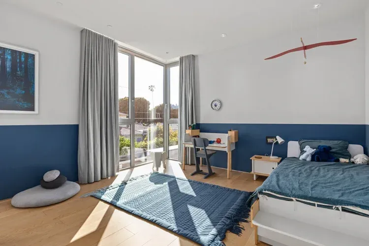 Bright bedroom with blue-and-white walls, bed, desk, rug, and sunlight from balcony doors