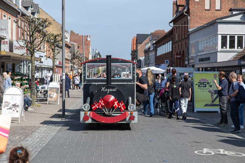 Et sort turisttog dekoreret med flag kører ned ad en solrig gade omkranset af bygninger og mennesker.