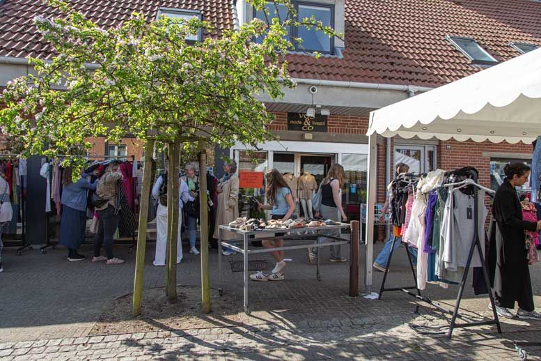Gademarked med tøjstativer og kunder uden for en murstensbygning på en solskinsdag.