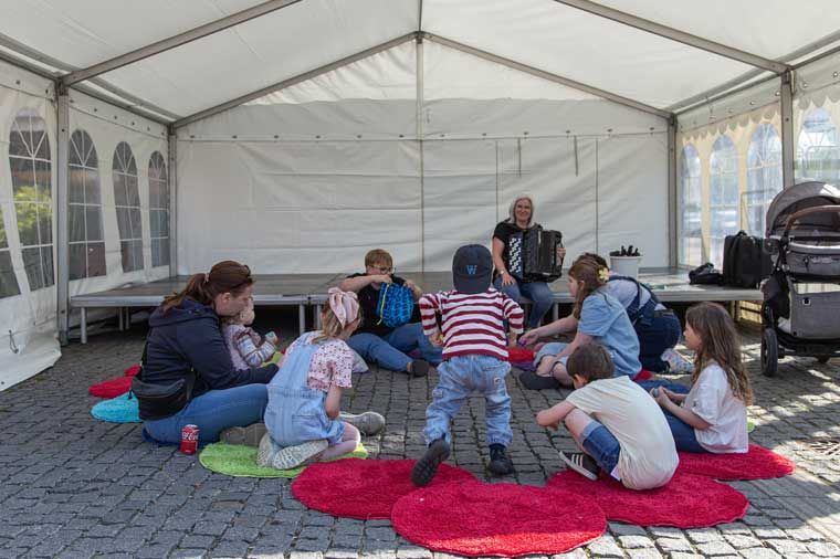 Børn og voksne sidder i et telt og lytter til en musiker, der spiller harmonika.