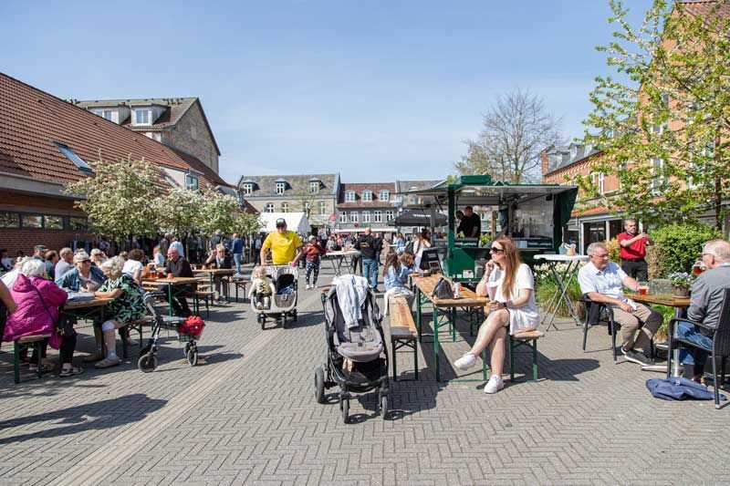 Udendørs markedsscene med folk ved borde, madvogn, barnevogne, solskinsdag.