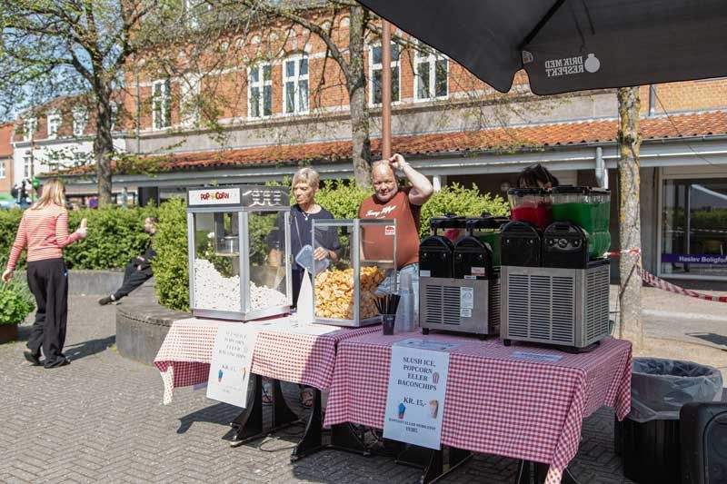 Popcorn- og isbod udendørs. To personer betjener kunder. Rød og hvid ternet dug.