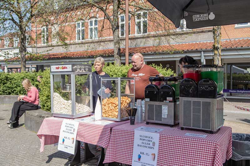 Popcorn- og drinksbod ved et udendørs arrangement; to personer bag disken, ternet dug, solskinsdag.