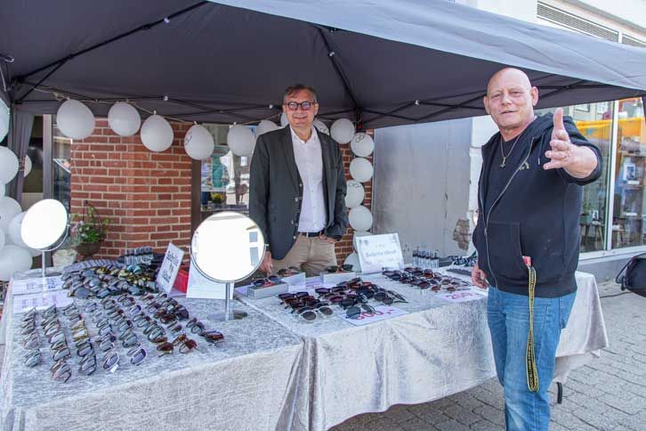 To mænd står med en solbrilleudstilling under en baldakin; den ene peger mod kameraet. Udendørs omgivelser med balloner.