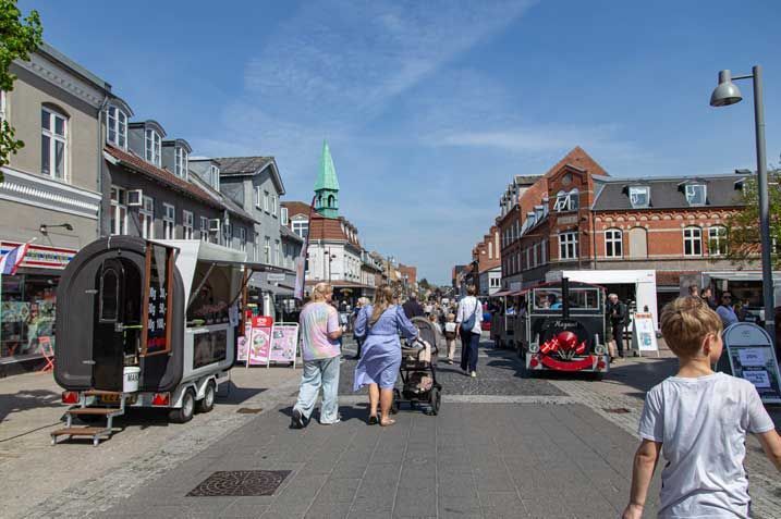 Gadebillede med butikker, madvogne og fodgængere under en solrig blå himmel.