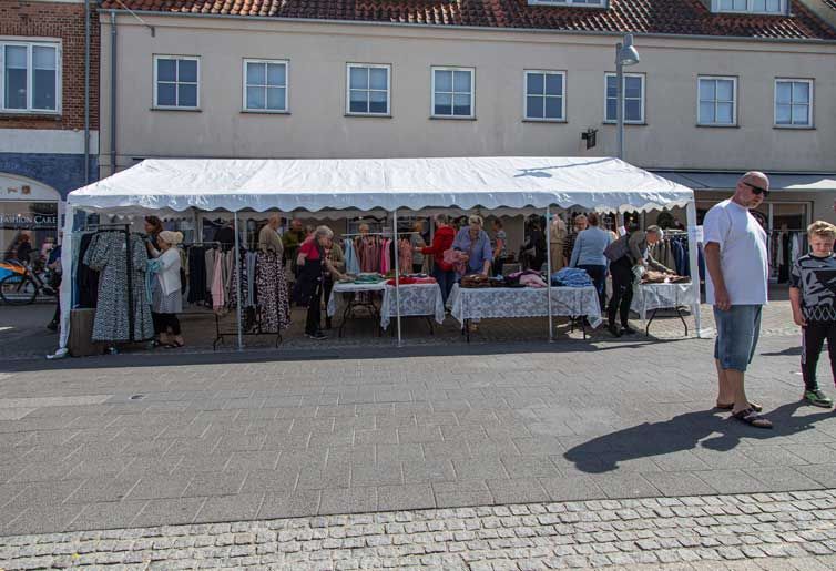 Folk kigger på tøj på et udendørs marked under et hvidt telt på en solskinsdag.