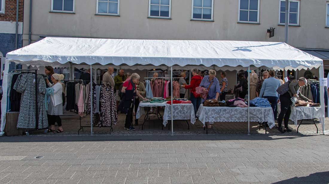 Folk kigger på tøj, der er udstillet på borde og hylder under et hvidt telt; en butiksfacade er baggrunden.