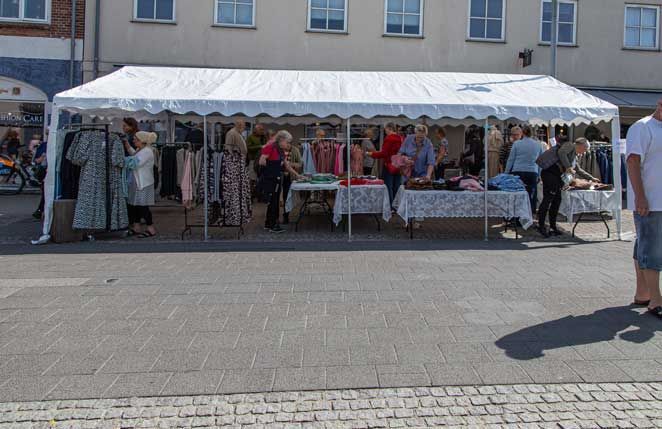 Hvidt telt med tøj til salg, folk kigger på borde udenfor, solskinsdag.