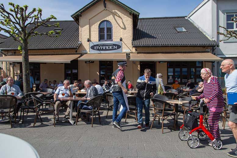 Folk ved udendørs caféborde; bygning med markise. Ældre person med rollator.