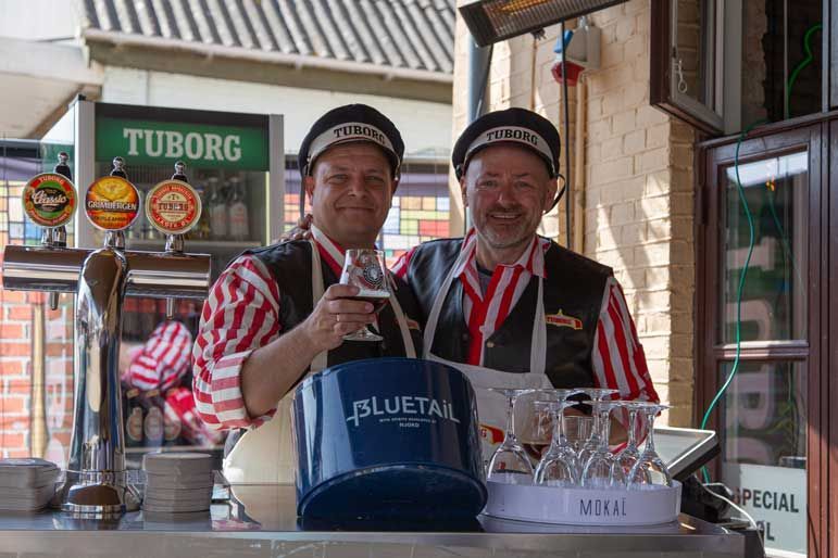 To mænd i matchende uniformer holder drinks i hånden på en udendørs bar. Tuborg-ølskilt i baggrunden.