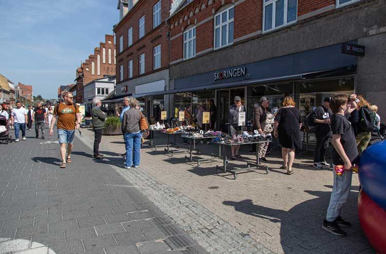 Folk kigger på varer, der er udstillet på borde uden for en butik på en solrig gade.