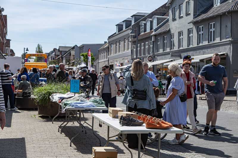 Udendørs marked på en gade. Folk kigger på varer ved bordet og går forbi bygninger.