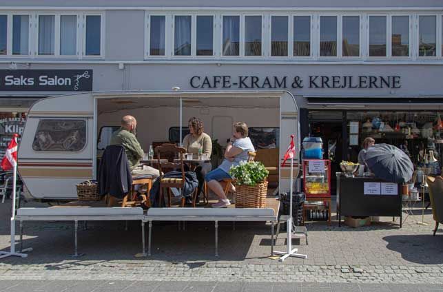 Cafe-Kram & Krejlerne café med folk siddende uden for en campingvogn; en butik og gade i baggrunden.
