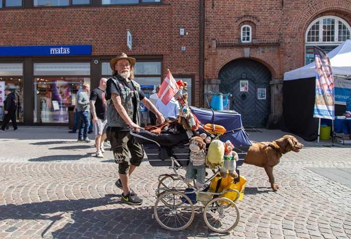 Mand med dukkefyldt klapvogn og hund på murstensgade, butik og buegang i baggrunden, solskinsdag.