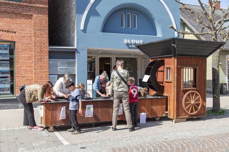 Folk ved en træbod med et vandhjul foran en blå bygning med en buet facade.