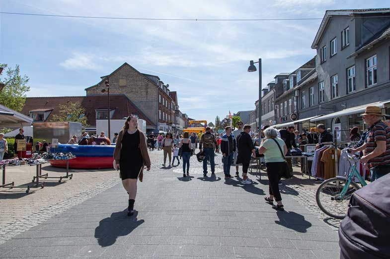 Gademarkedscene; folk går og kigger på boder med varer under en blå himmel, bygninger i baggrunden.