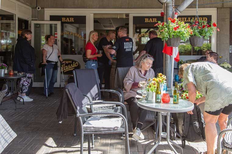 Folk uden for en bar med borde og stole. Nogle drikker, andre samtaler. En urtepotte hænger ovenover.