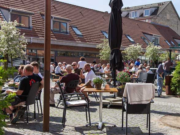 Udendørs caféscene med folk siddende ved borde på en solskinsdag. Bygninger med røde tegltage i baggrunden.