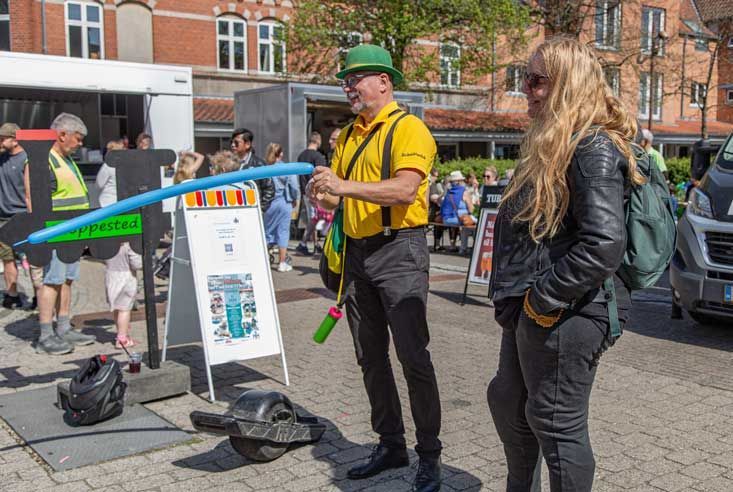 En ballonkunstner i en gul skjorte og grøn hat laver en ballonkreation på en offentlig plads.