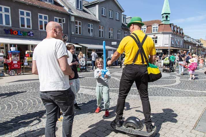 Person på ethjulet køretøj giver et ballonsværd til et barn på en trafikeret gade. Bygninger i baggrunden.