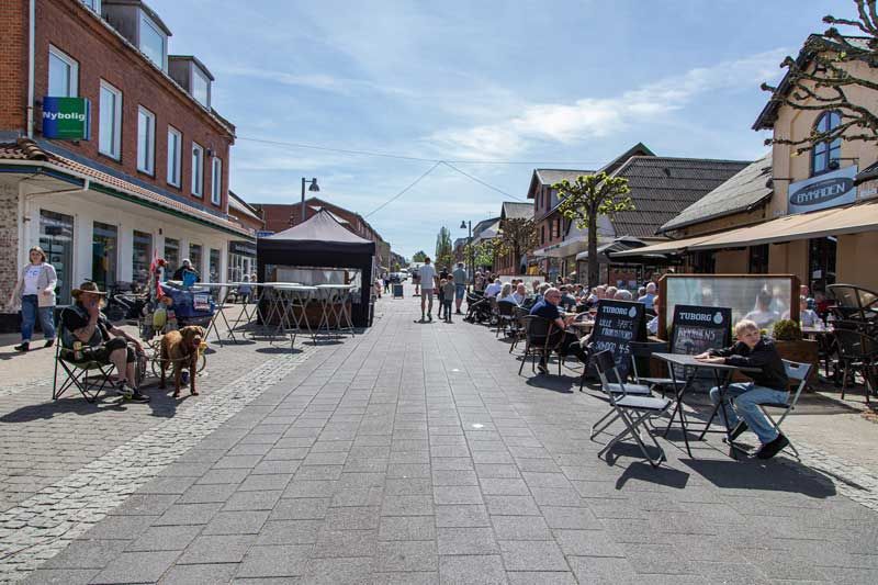 Brostensbelagt gade med udendørs siddepladser og butikker på en solskinsdag. Folk spiser og går tur.