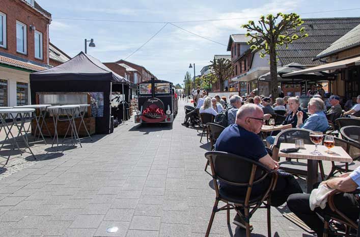 Gadescene med udendørs siddepladser, madsælger og et lille køretøj på den brostensbelagte vej på en solskinsdag.