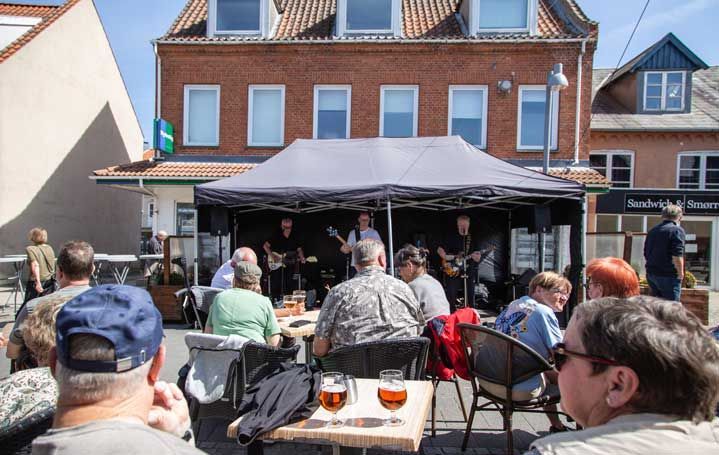 Band optræder på udendørs scene, publikum siddende ved borde med drinks. Murstensbygning i baggrunden.