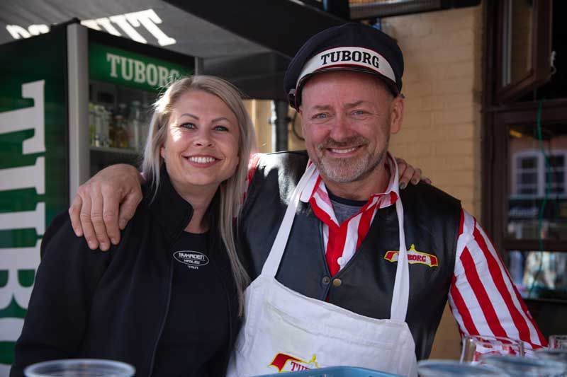 Kvinde og mand med Tuborg-skilte, smilende. Mand i stribet forklæde og hat.