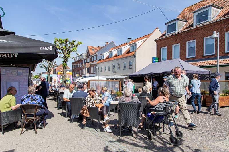 Folk sidder ved udendørs borde på en solskinsdag. Bygninger og et telt er i baggrunden.