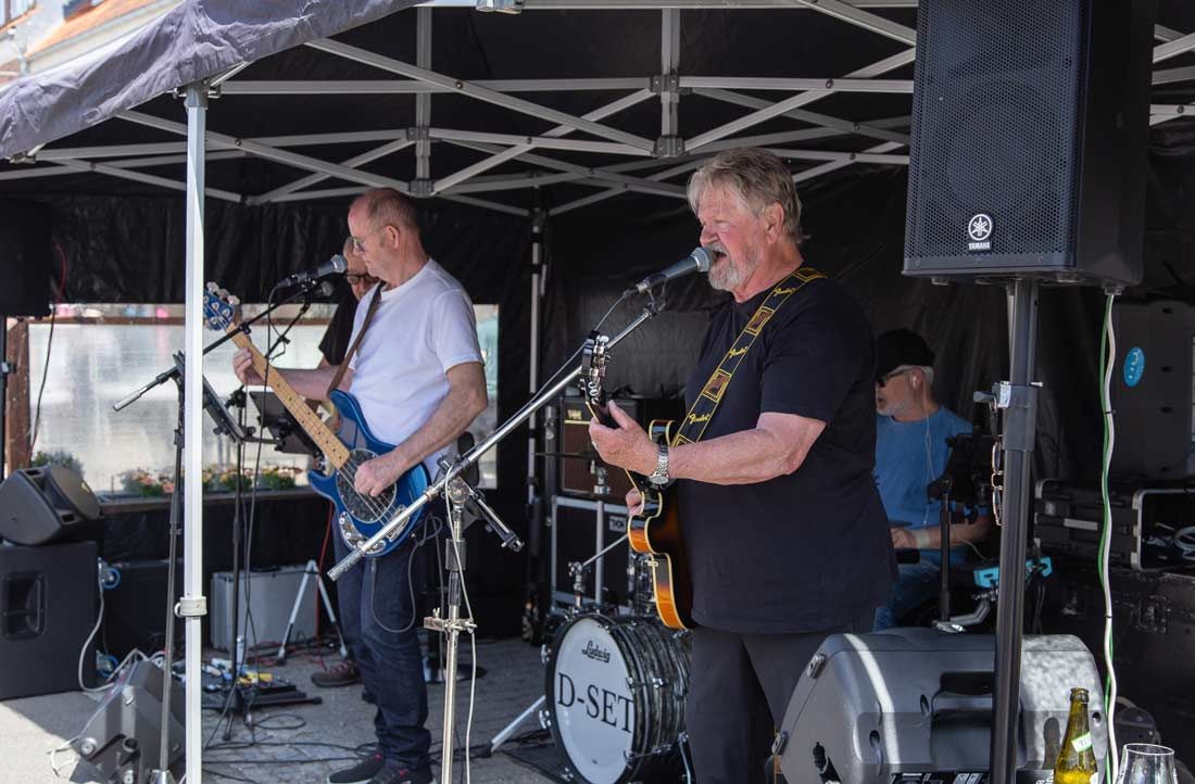 Bandet optræder live på en scene udendørs under et telt. En guitarist synger. En anden spiller bas. Trommer i baggrunden.