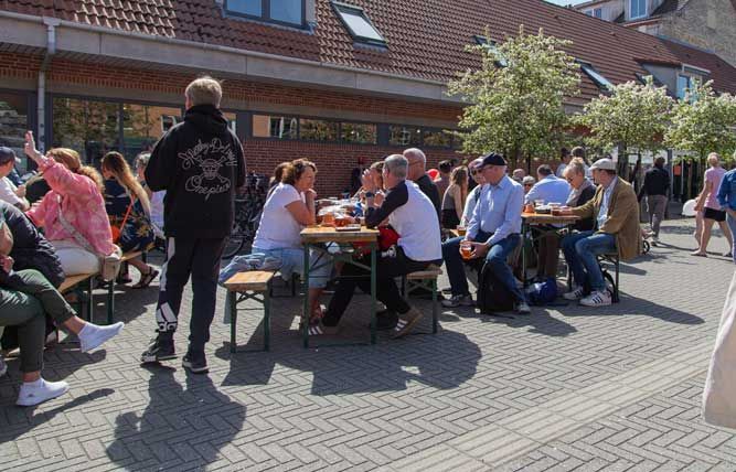Folk sidder ved udendørs borde, drikker og snakker på en solrig gade, nær bygninger.