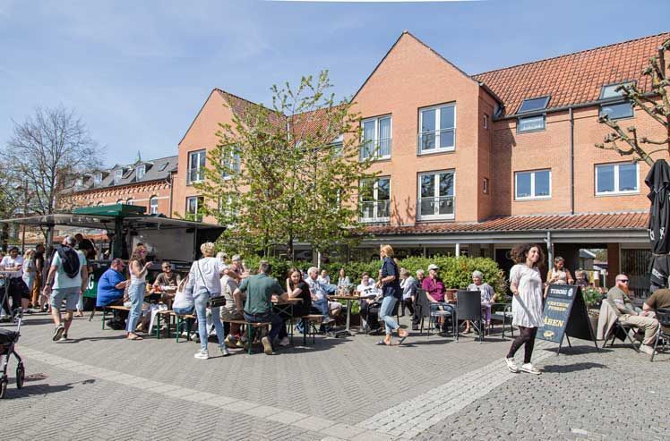 Udendørs café med folk siddende ved borde, rød murstensbygning i baggrunden. Solskinsdag.