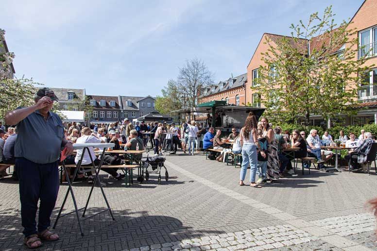 Udendørs sammenkomst med folk ved borde. En mand justerer glassene i forgrunden. Bygninger og en foodtruck i baggrunden.
