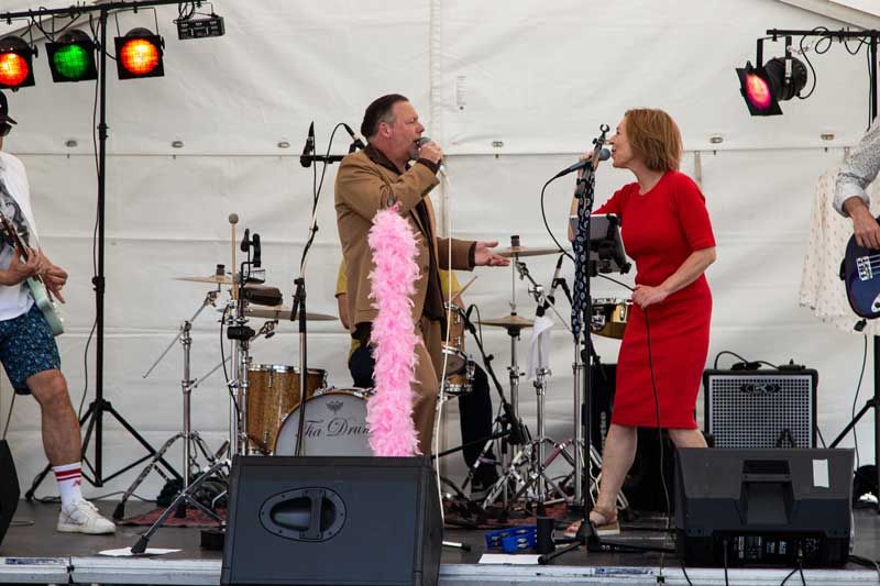 Bandet optræder på scenen under et hvidt telt. Mand i brun jakke og kvinde i rød kjole synger ind i mikrofonerne.