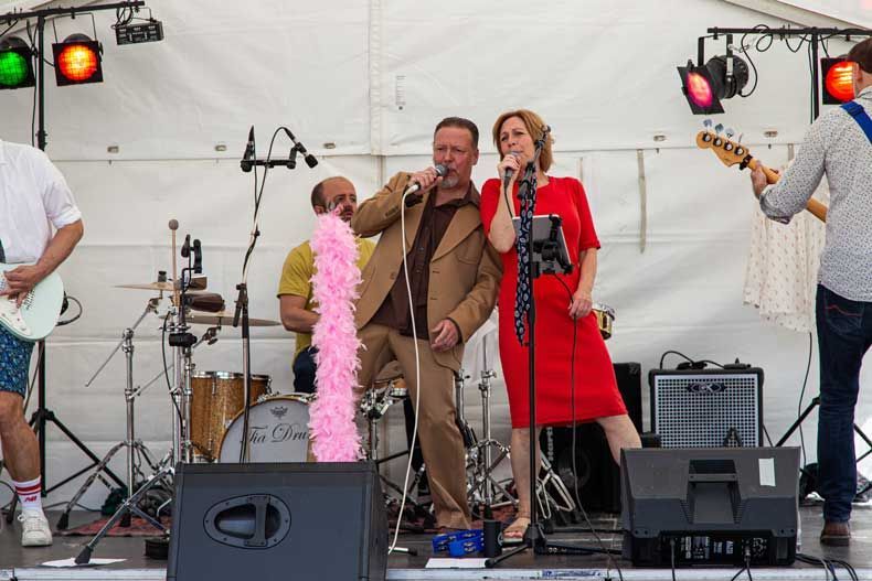 Band optræder på scenen under et telt. To sangere med mikrofoner, flankeret af musikere og en lyserød boa.