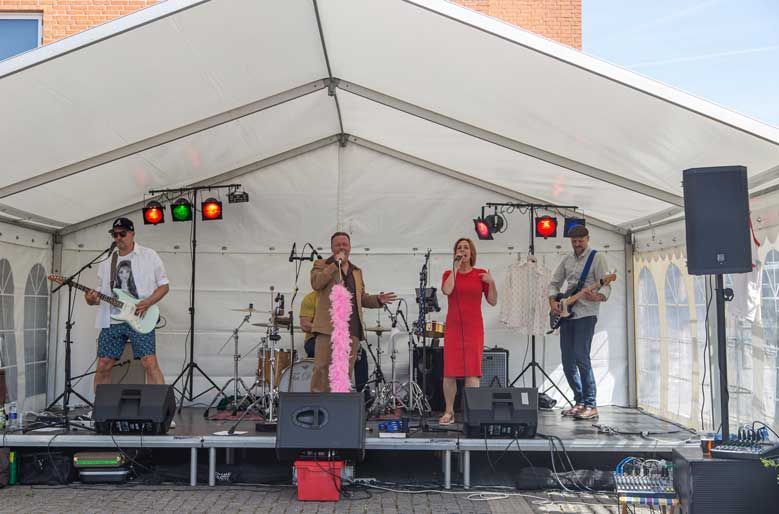 Band optræder på scenen under et hvidt telt; sanger i rød kjole, guitarist, trommeslager og bassist.